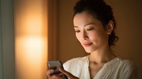 Young Asian woman in white sweater using smartphone at home with warm evening lighting creating cozy atmosphere for technology and lifestyle content.