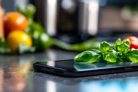 Fresh basil leaves on black smartphone screen with colorful tomatoes in background, modern food photography composition on dark kitchen counter.