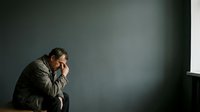 Mature man with gray hair and stubble, wearing olive jacket, sitting in contemplative pose with hand to face against neutral wall background, expressing stress, worry, or deep thought for business and lifestyle concepts.
