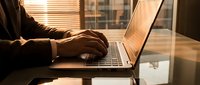 Professional businessman typing on laptop computer at modern office desk during golden hour sunset with warm lighting through window blinds.