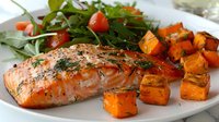 Grilled salmon fillet with roasted butternut squash cubes and fresh arugula salad garnished with cherry tomatoes and herbs, served on white plate.