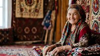 Senior woman of Central Asian ethnicity wearing traditional red embroidered clothing and headscarf, sitting on ornate patterned carpets and rugs in a textile workshop or home studio setting.