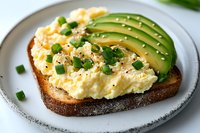 Creamy scrambled eggs on toasted dark bread garnished with sliced avocado, fresh chives and black pepper served on white ceramic plate for breakfast.
