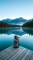 Photographer sitting on wooden dock capturing mountain lake landscape with camera, serene alpine scenery with mirror water reflection and evergreen forest for travel and nature content.