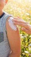 Woman applying sunscreen lotion to arm outdoors in sunny field with blooming flowers, protecting skin from UV rays and sun damage during summer.