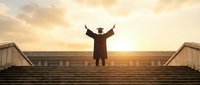 Silhouette of businessman in suit standing on steps with raised arms celebrating success against golden sunset sky. Achievement and victory concept.