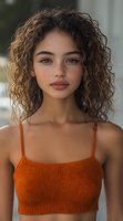 Young mixed race woman with natural curly hair and freckles wearing orange knit crop top, looking directly at camera with soft natural makeup and confident expression.