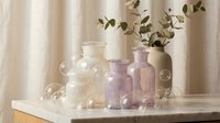 Elegant glass bottles and vases in soft pastel tones arranged on wooden tray with eucalyptus branches creating minimalist home decor styling composition.