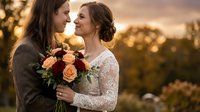 Two brides in elegant wedding dresses share intimate moment during golden hour sunset ceremony with bridal bouquet of roses and greenery.