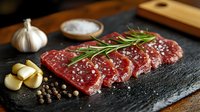 Raw beef steak slices seasoned with salt, rosemary, and spices on dark slate board with garlic cloves and peppercorns, ready for cooking.