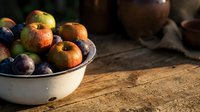 Assorted fresh apples and plums in white enamel bowl on rustic wooden surface with dark background, suitable for food styling and culinary projects.