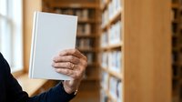 Senior man holding blank white book in library with wooden shelves, suitable for publishing, education, and literary projects.