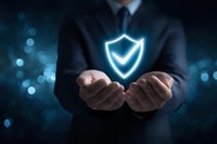 Businessman holding glowing shield symbol representing cybersecurity, data protection, and digital safety in corporate environment against blue bokeh background.