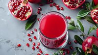 Fresh pomegranate juice in glass with ripe cut fruits, scattered seeds and green leaves on gray concrete background, healthy antioxidant drink.