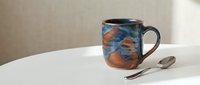 Handcrafted ceramic mug with blue and terracotta glaze patterns sits on white table with silver spoon, perfect for coffee tea and kitchen photography.