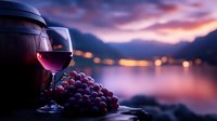 Red wine glass and fresh grapes against blurred evening cityscape with dramatic purple sunset sky and bokeh lights reflecting on calm water surface.