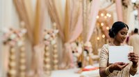 Young South Asian woman in traditional gold embroidered attire holding blank card at elegant wedding venue with floral decorations and warm lighting for celebrations.