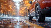 Orange crossover SUV on wet autumn road with fallen leaves, low angle view focusing on wheel and tire against blurred fall foliage background in golden colors.