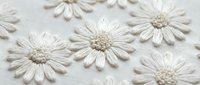 Delicate white daisy flowers embroidered on cream fabric with intricate stitching details. Handcrafted textile art for fashion and home decor projects.