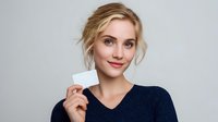 Young blonde woman in navy sweater holding blank white card against gray background, showcasing casual professional style for business or identity mockup.