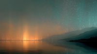 Northern lights aurora borealis dancing over calm lake with mountain silhouettes and golden sunset glow reflecting on water surface creating magical atmosphere.