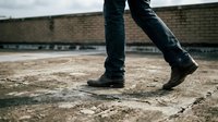 Man walking in worn leather boots and blue jeans on a rough urban rooftop surface with a brick wall background.