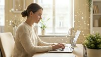Young Caucasian woman in beige sweater working on laptop at home office desk by window with warm natural lighting and cozy atmosphere for remote work.
