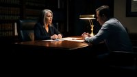 Female lawyer with gray hair in dark blazer conducting client consultation across wooden desk in dimly lit office with law books and desk lamp, professional legal meeting.