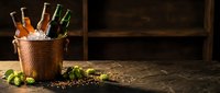 Copper bucket filled with assorted beer bottles on ice, hops and spices scattered on rustic wooden surface for brewery advertising and beverage marketing.