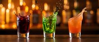 Three colorful cocktails with splashing liquid in glasses on bar counter with warm bokeh lights background for beverage menu design.
