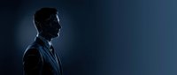 Silhouette of a professional man in business suit and tie standing in profile against dark blue background with dramatic lighting for corporate and leadership concepts.