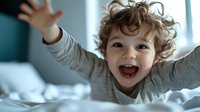Joyful curly-haired child with arms raised, laughing and playing on bed in morning light. Natural lifestyle portrait capturing pure happiness and carefree childhood moments.
