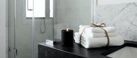 Rolled white towels with twine on black bathroom counter beside black candle, modern shower enclosure and marble wall backdrop.