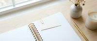 Minimalist workspace with blank notebook, envelope and wooden pencils on natural wood desk near window for creative writing and correspondence.
