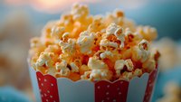 Fresh buttery popcorn overflowing from striped red and white container against blurred background, macro shot with selective focus for entertainment and snack concepts.