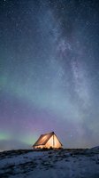 Illuminated camping tent under spectacular Milky Way galaxy with aurora borealis lights in starry night sky over snowy winter landscape for adventure travel.