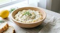 Creamy hummus dip with olive oil and herbs served in wooden bowl with fresh lemon and bread for appetizer or snack preparation.