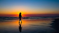 Surfer holding board walking along beach at golden sunset, silhouetted against ocean waves and colorful sky, perfect for travel and lifestyle content.