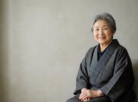 Senior Asian woman with gray hair wearing dark traditional kimono, sitting with hands folded, neutral background, suitable for healthcare, wellness, cultural diversity, and lifestyle editorial use.