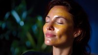 Young woman with closed eyes and glowing gold glitter makeup on face against dark background with bokeh lights, expressing serenity and wellness.