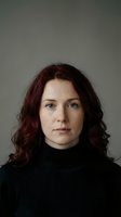 Professional portrait of confident Caucasian woman with auburn curly hair wearing black turtleneck sweater against neutral gray background.
