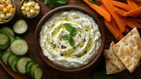 Creamy herb dip with olive oil drizzle in rustic bowl, served with fresh cucumber slices, carrot sticks, crackers and cheese cubes on wooden table.