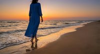 Woman in blue dress walking on beach at sunset with golden sky and ocean waves creating peaceful vacation atmosphere for travel and wellness concepts.