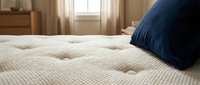 Comfortable beige quilted mattress with navy blue pillow in bright bedroom with window and wooden furniture for sleep and relaxation.