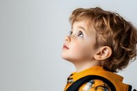 Young caucasian child with curly hair wearing orange and plaid clothing looking up with wonder and curiosity against light background, profile view portrait.