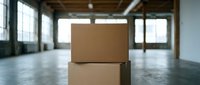 Stacked cardboard boxes in empty warehouse with natural light from windows, suitable for logistics, shipping, storage and delivery concepts.