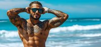 Tattooed muscular man with sunglasses posing shirtless on tropical beach with turquoise ocean waves. Summer vacation lifestyle and fitness concept.