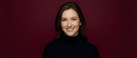 Professional businesswoman with dark hair smiling confidently in black turtleneck sweater against burgundy background for corporate headshots and marketing.