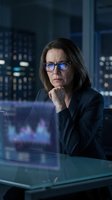 Middle-aged woman with dark hair wearing glasses and black blazer, sitting at desk with holographic data display in modern office with city skyline view for business and technology concepts.