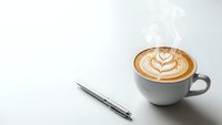 Steaming hot cappuccino with latte art heart pattern next to silver pen on clean white surface for morning productivity and cafe workspace concepts.